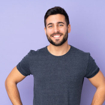 Caucasian handsome man posing with arms at hip and smiling over isolated purple background