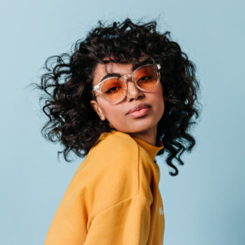 Dreamy young woman in sunglasses looking at camera. Front view of interested curly girl isolated on blue background.