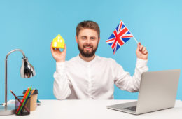 Self confident bearded man sitting at workplace with laptop, holding Great Britain flag and paper house, dreaming to buy accommodation in England. Indoor studio shot isolated on blue background.
